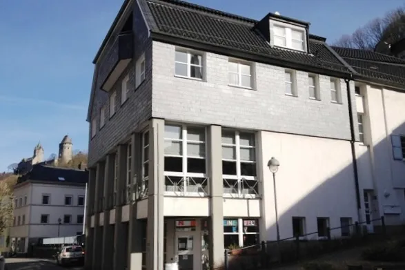 Das Lutherhaus in Altena mit grauer Schieferfassade und großen Fenstern vor einer Burg auf einem Hügel.