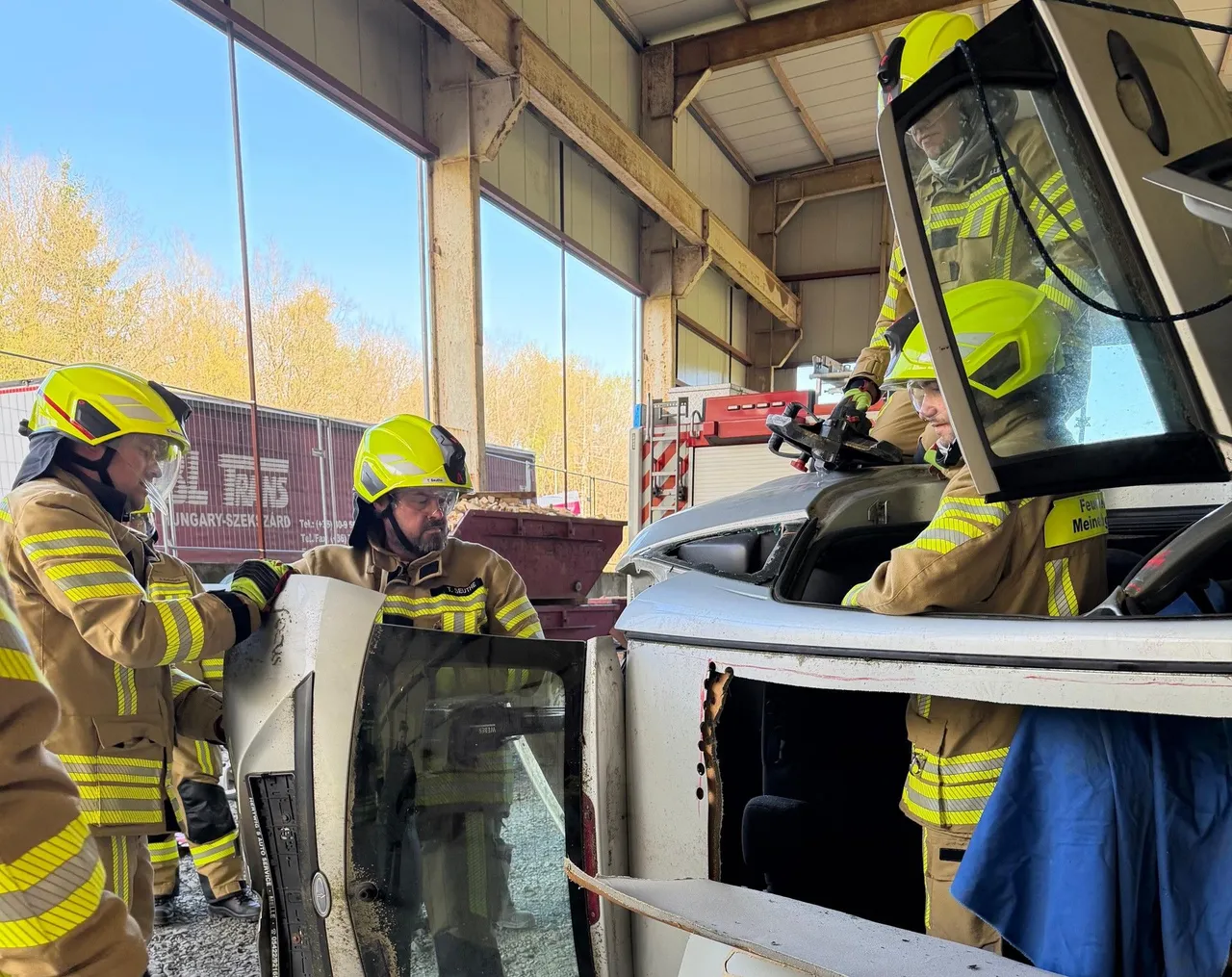 Drei Feuerwehrleute in voller Schutzausrüstung arbeiten an einem stark beschädigten Fahrzeug, während ein Kollege im Hintergrund das Geschehen beobachtet. Im Hintergrund ist ein LKW zu sehen.