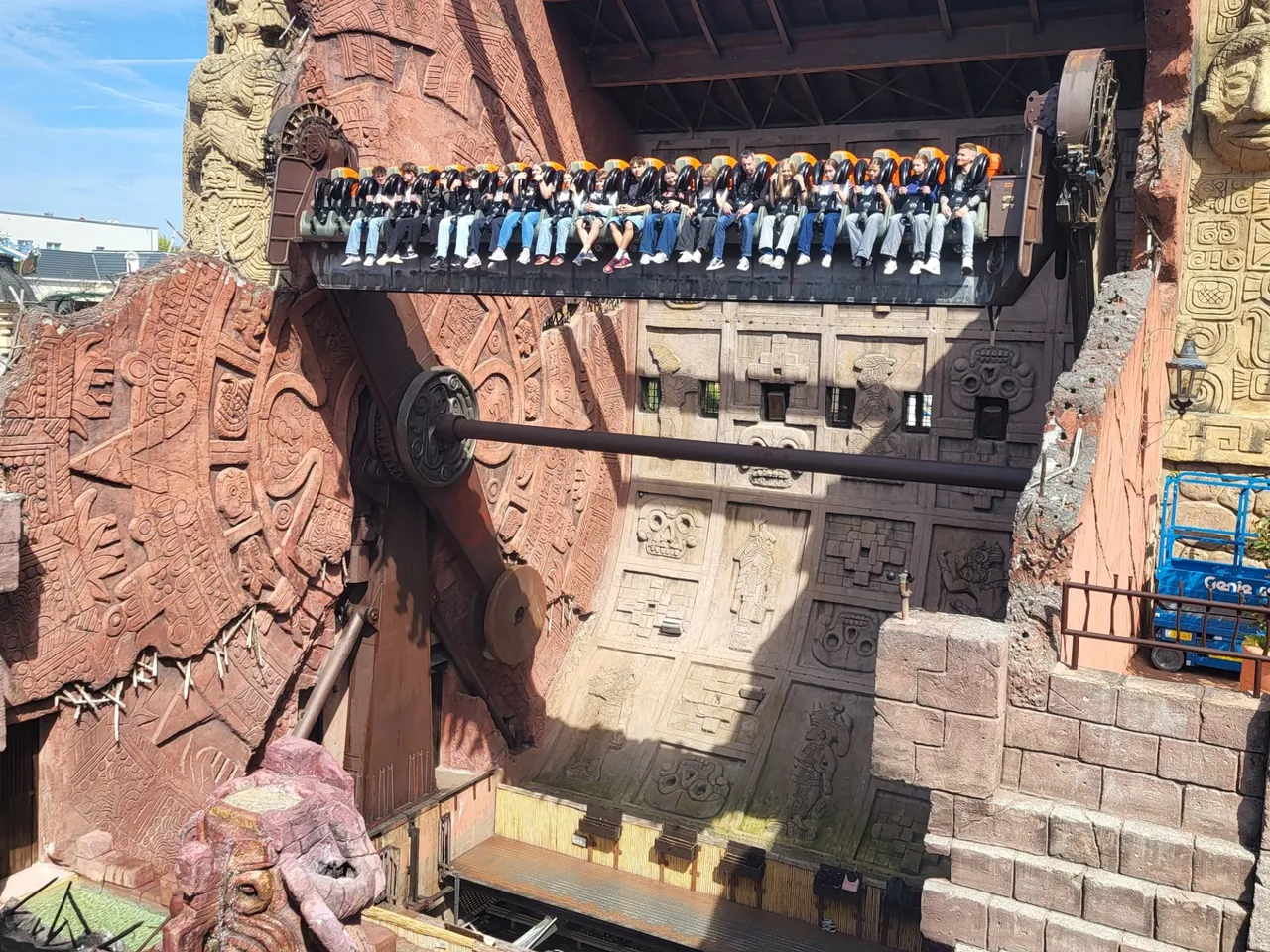 Eine Achterbahnfahrt mit vielen Fahrgästen vor einer detailreichen, tempelartigen Kulisse. Im Vordergrund ein Baugerüst und eine Hebebühne.