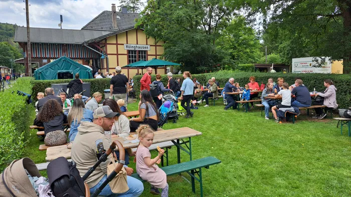 Menschen sitzen an Biertischgarnituren vor dem Bahnhof Hüinghausen und genießen den Tag. Im Hintergrund ist ein Zelt aufgebaut und ein Schild mit der Aufschrift „Hüinghausen“ zu sehen.
