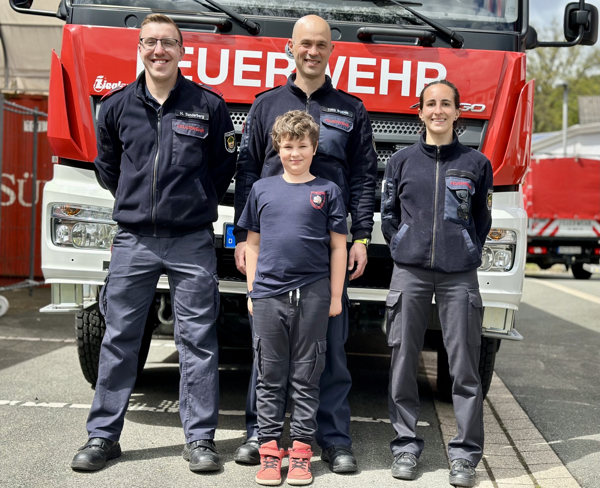 Vier Feuerwehrleute und ein Kind stehen vor einem roten Feuerwehrauto. Sie tragen Uniformen und lächeln in die Kamera.