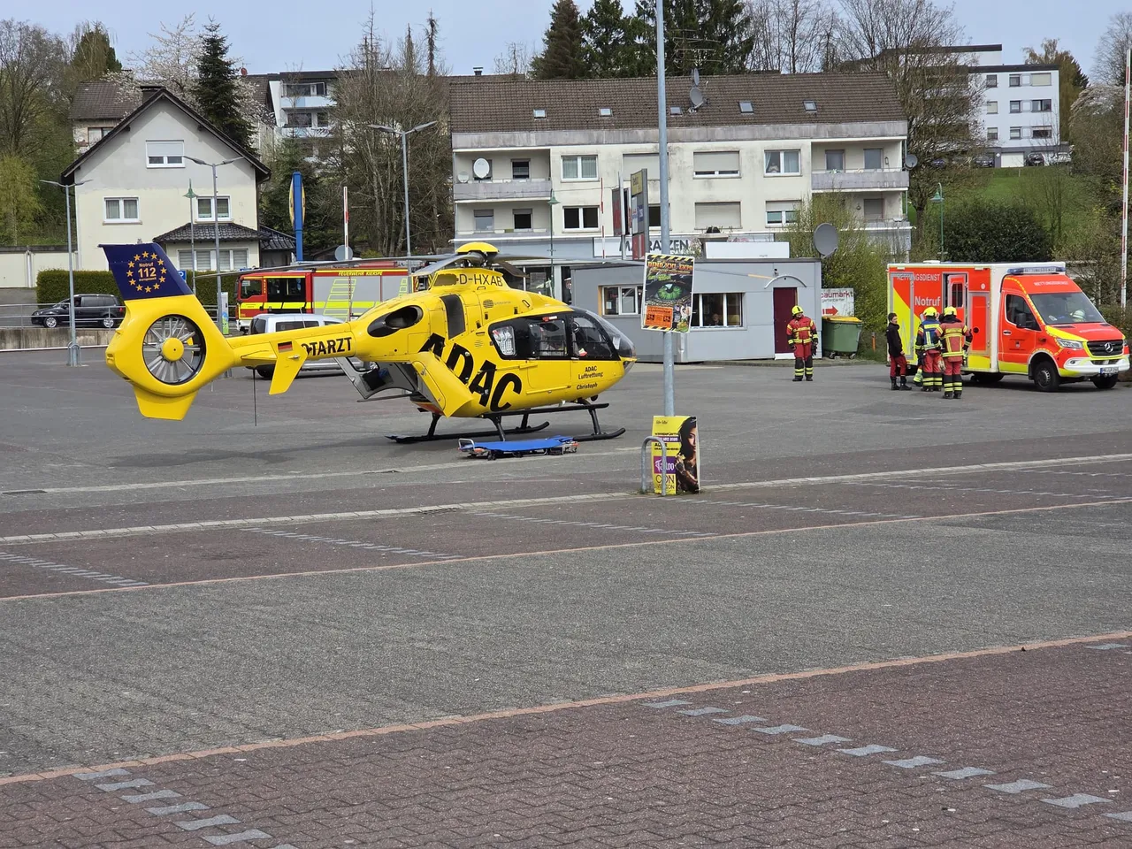Gelber Rettungshubschrauber mit dem Aufdruck ADAC und dem Kennzeichen D-HXAB landet auf einem gepflasterten Platz vor einem Einkaufszentrum, während Feuerwehrleute in der Nähe stehen.