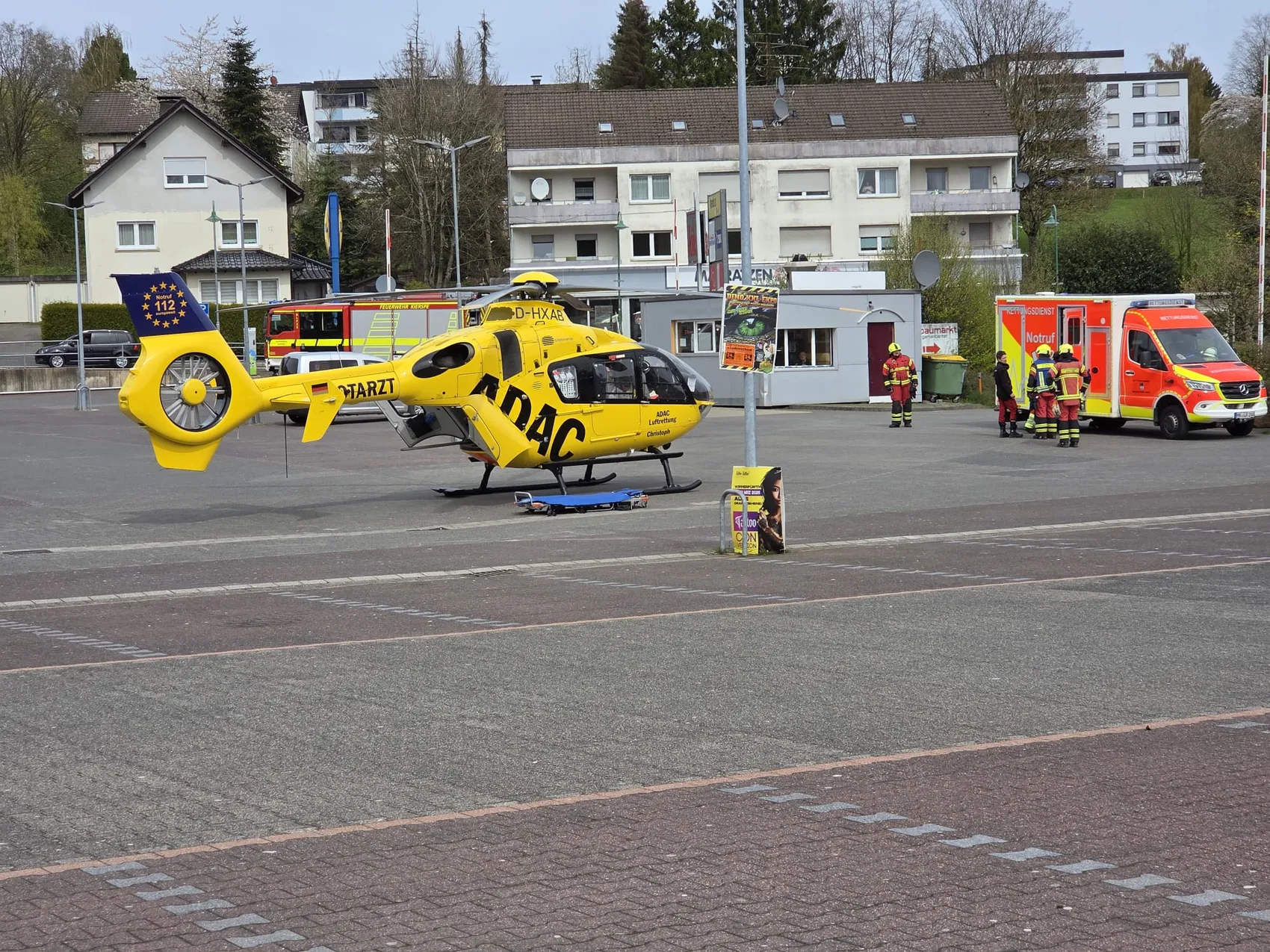 Gelber Rettungshubschrauber mit dem Aufdruck ADAC und dem Kennzeichen D-HXAB landet auf einem gepflasterten Platz vor einem Einkaufszentrum, während Feuerwehrleute in der Nähe stehen.