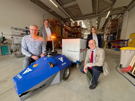 Four people stand in a warehouse with a small blue robot in the foreground and a larger white robot behind them. From left to right are Nico Hackmann, Jochen Schröder, Sonja Pfaff, and Landrat Ralf Schwarzkopf.