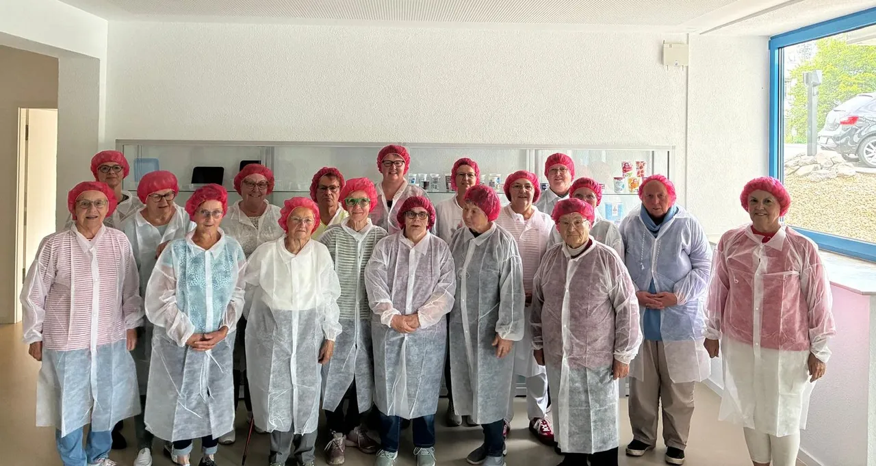 Eine Gruppe von etwa zwanzig älteren Frauen in Schutzhauben und weißen Kitteln steht in einem hellen Raum vor Regalen mit Produkten. Sie besuchen offenbar die Firma Fernholz.