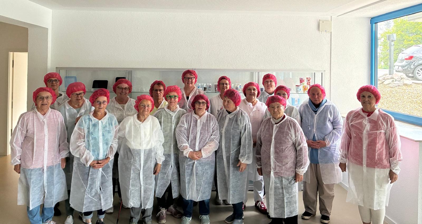 Eine Gruppe von etwa zwanzig älteren Frauen in Schutzhauben und weißen Kitteln steht in einem hellen Raum vor Regalen mit Produkten. Sie besuchen offenbar die Firma Fernholz.