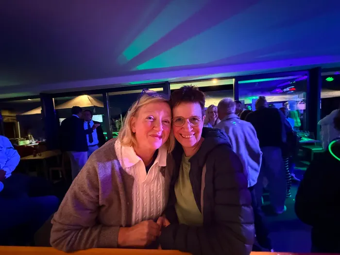 Zwei lächelnde Frauen posieren bei einer Abendveranstaltung im Tennisclub unter farbiger Beleuchtung.