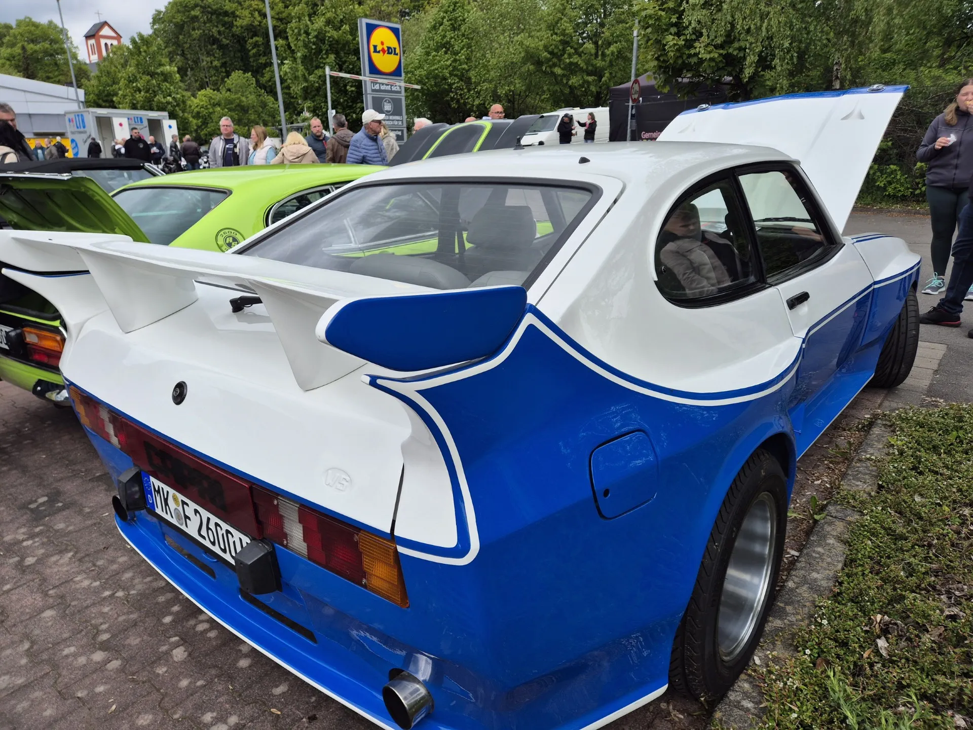 Heckansicht eines weißen Oldtimers mit blauen Streifen und großem Heckspoiler, Kennzeichen MV-F 2600. Im Hintergrund sind weitere Oldtimer und Besucher zu sehen.