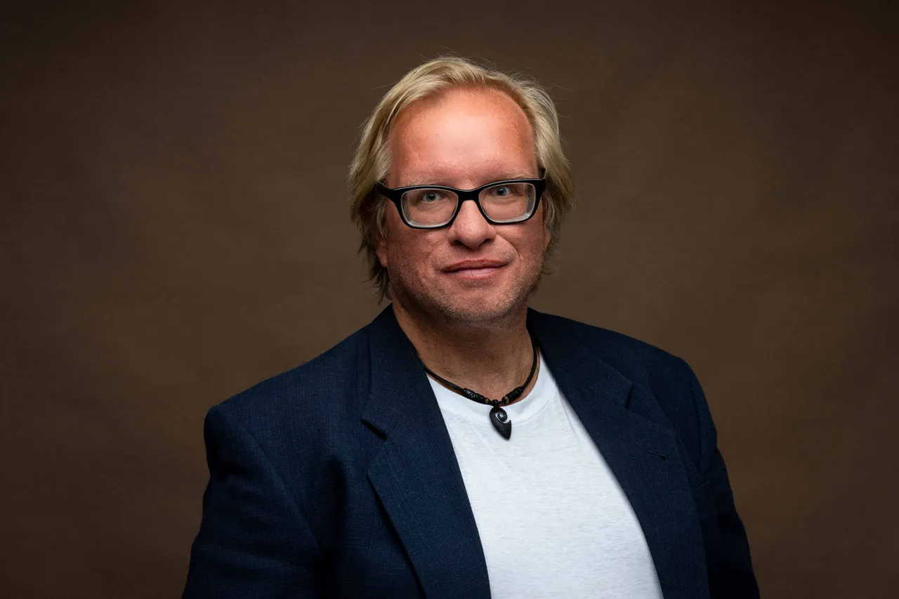 Thomas Höller mit Brille, blauem Sakko und weißem T-Shirt vor einem braunen Hintergrund.