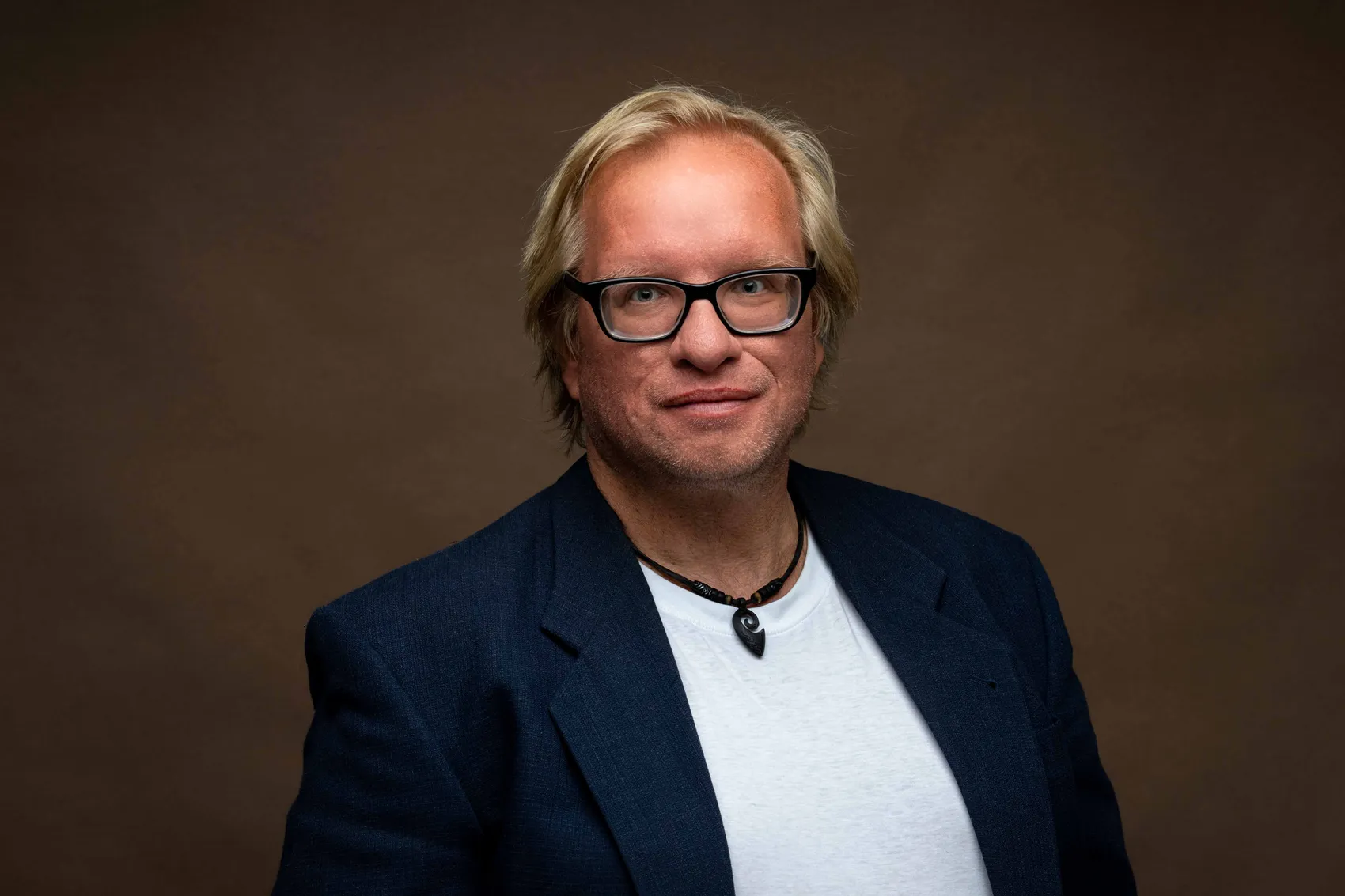 Thomas Höller mit Brille, blauem Sakko und weißem T-Shirt vor einem braunen Hintergrund.