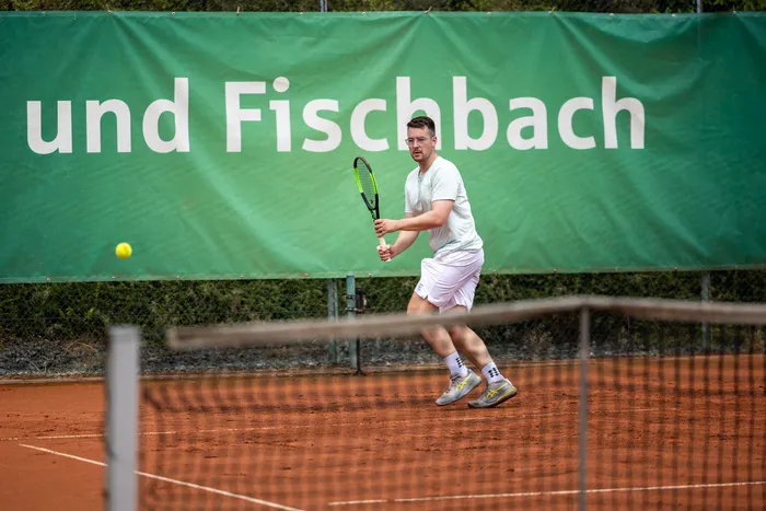 Ein Tennisspieler in weißer Kleidung bereitet sich auf einem Sandplatz darauf vor, einen gelben Ball zu schlagen, vor einem grünen Banner.