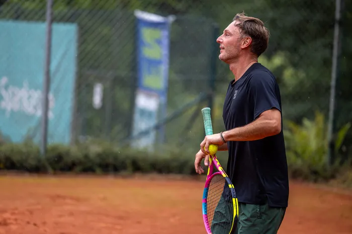 Ein Mann in schwarzem T-Shirt steht auf einem Sandplatz und hält einen bunten Tennisschläger sowie einen gelben Ball.