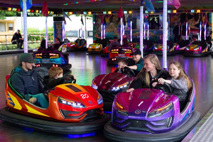 Menschen jeden Alters fahren auf der Kirmes mit Autoscootern in bunten Fahrzeugen. Im Hintergrund sind weitere Fahrgeschäfte und Besucher zu sehen.