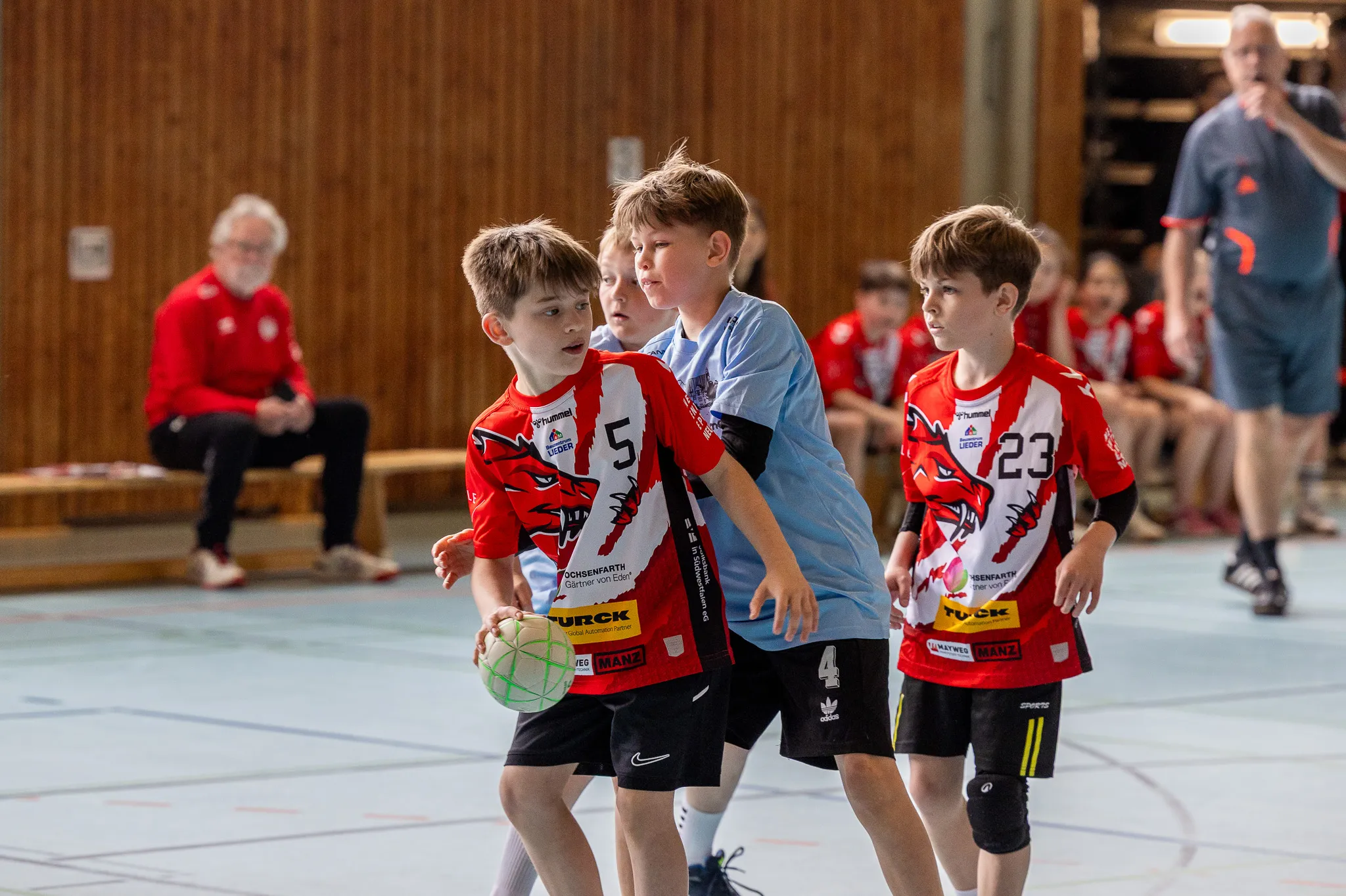 Junge Handballspieler in roten Trikots mit der Nummer 5, 4 und 23 trainieren in einer Sporthalle.