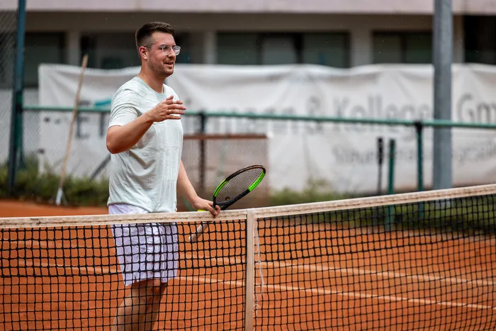 Ein Mann in weißer Sportkleidung steht mit einem Tennisschläger auf einem Sandplatz neben dem Netz.