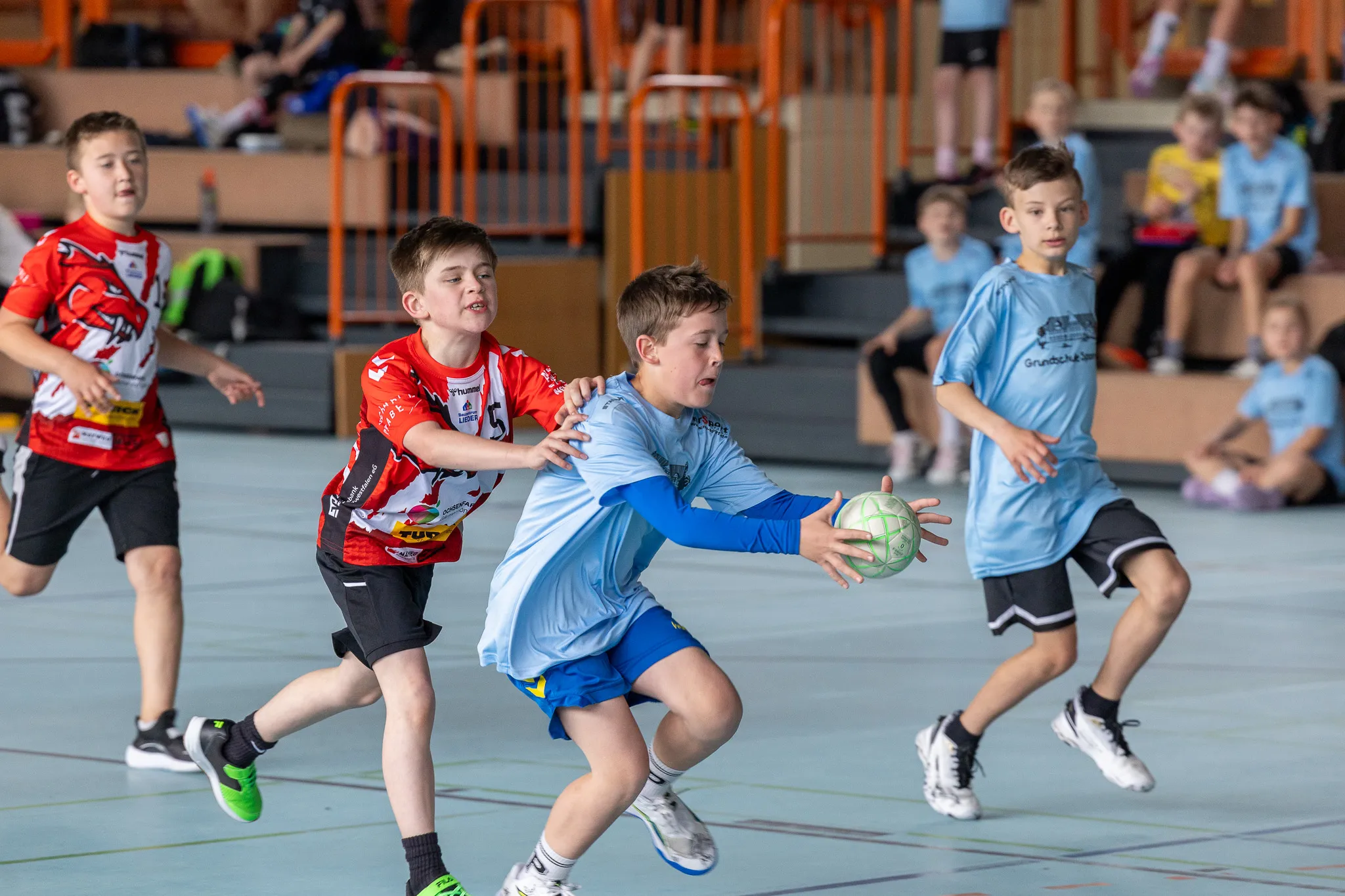 Zwei Handballspieler, einer in rotem Trikot, der andere in blauem Trikot, kämpfen um den Ball in einer Sporthalle.