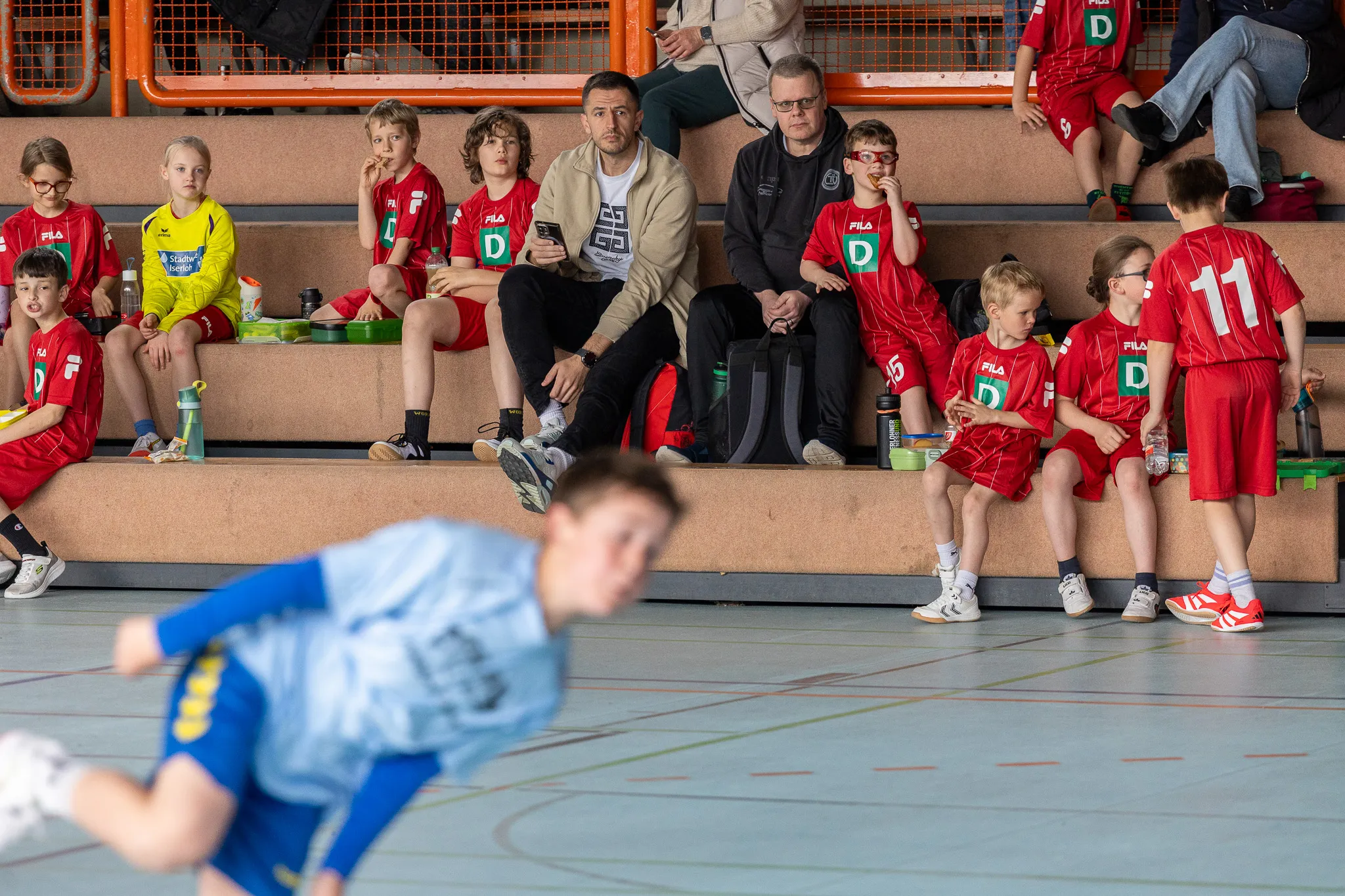 Mannschaft mit Trainer sitzt auf einer Stufe, während ein Spieler im Vordergrund einen Wurf vorbereitet.