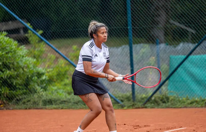Eine Tennisspielerin in weißem T-Shirt und schwarzem Rock steht mit einem roten Schläger bereit auf einem Sandplatz.