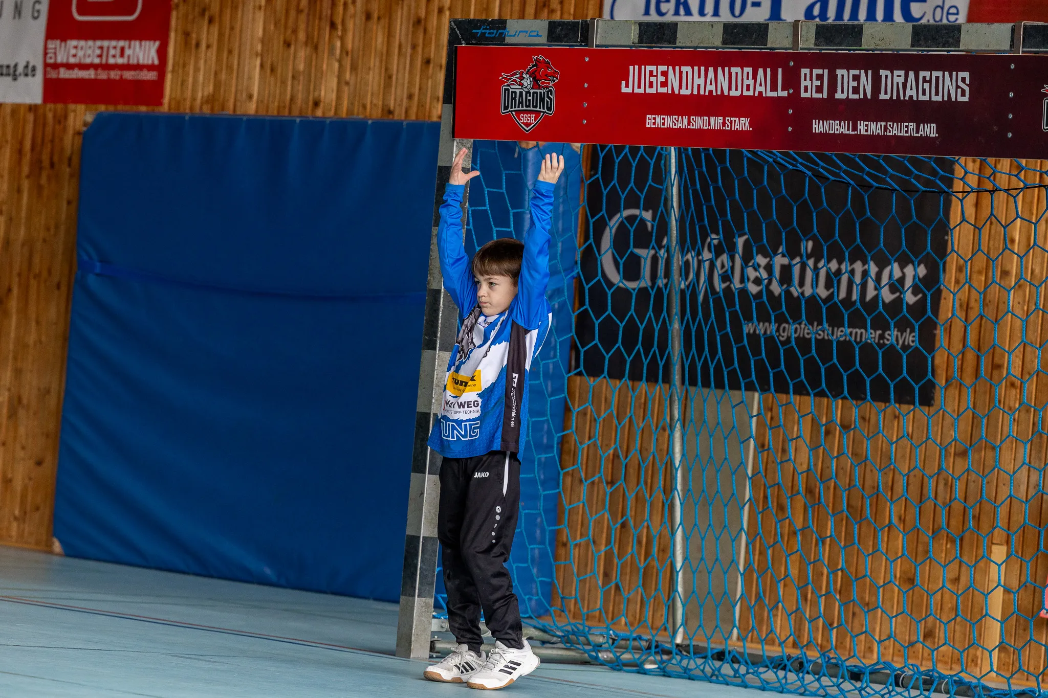 Ein Jugendhandballspieler mit erhobener Hand steht auf einem blauen Hallenboden vor einer Werbebande und einem Drahtzaun.