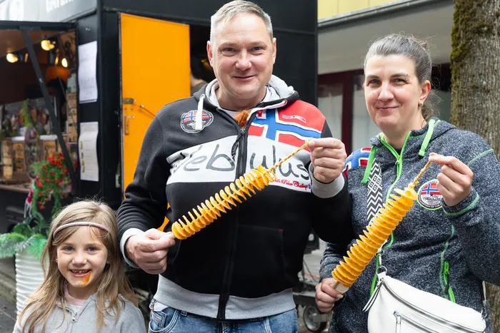 Ein Mann und eine Frau halten Spiral-Kartoffelchips vor einer jungen Frau. Im Hintergrund ein Kirmesstand.