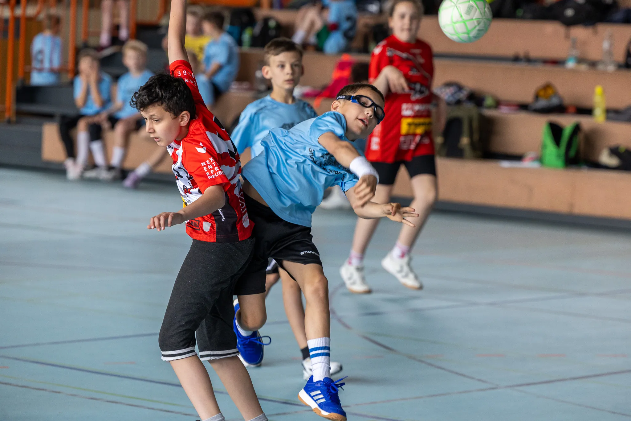 Zwei Handballspieler, einer im roten Trikot und einer im blauen Trikot, kämpfen um den Ball während eines Spiels in einer Sporthalle.