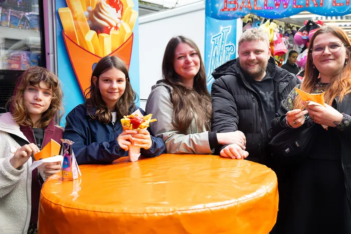 Fünf Personen lächeln in die Kamera, während sie vor einem Imbissstand auf der Kirmes stehen und Pommes oder Papierstücke in der Hand halten.