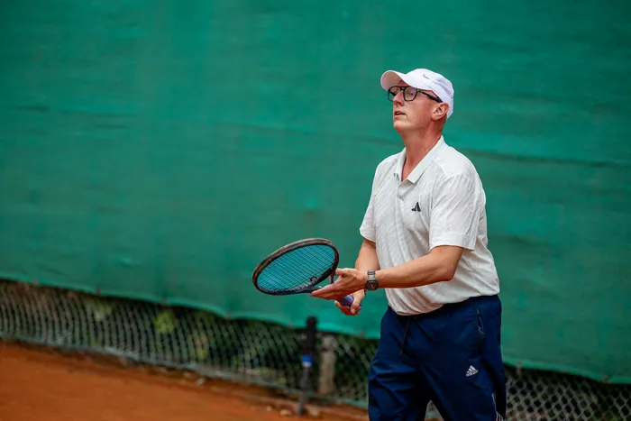 Ein Mann in weißem Poloshirt und weißer Kappe hält einen Tennisschläger und blickt auf einem Sandplatz nach oben.