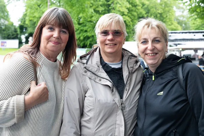 Drei lächelnde Frauen posieren eng nebeneinander für ein Foto. Im Hintergrund sind Bäume und Menschen zu sehen.