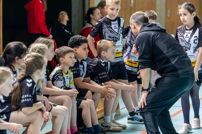 Eine Gruppe von Kindern in Kampfsportkleidung sitzt und steht um einen Trainer in einer Sporthalle.