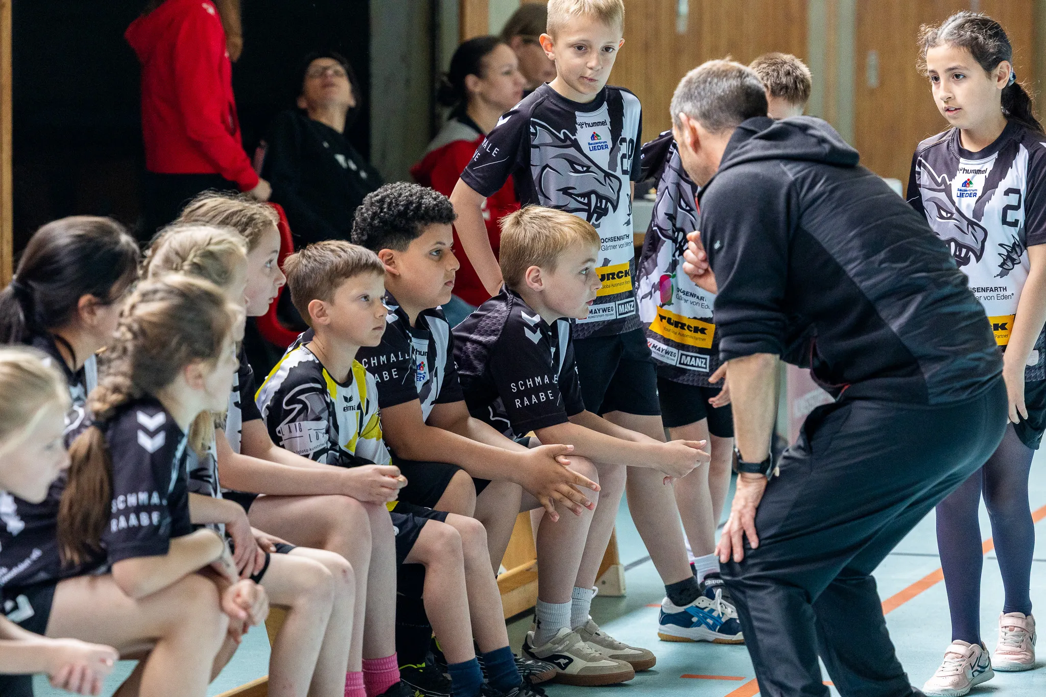 Eine Gruppe von Kindern in Kampfsportkleidung sitzt und steht um einen Trainer in einer Sporthalle.