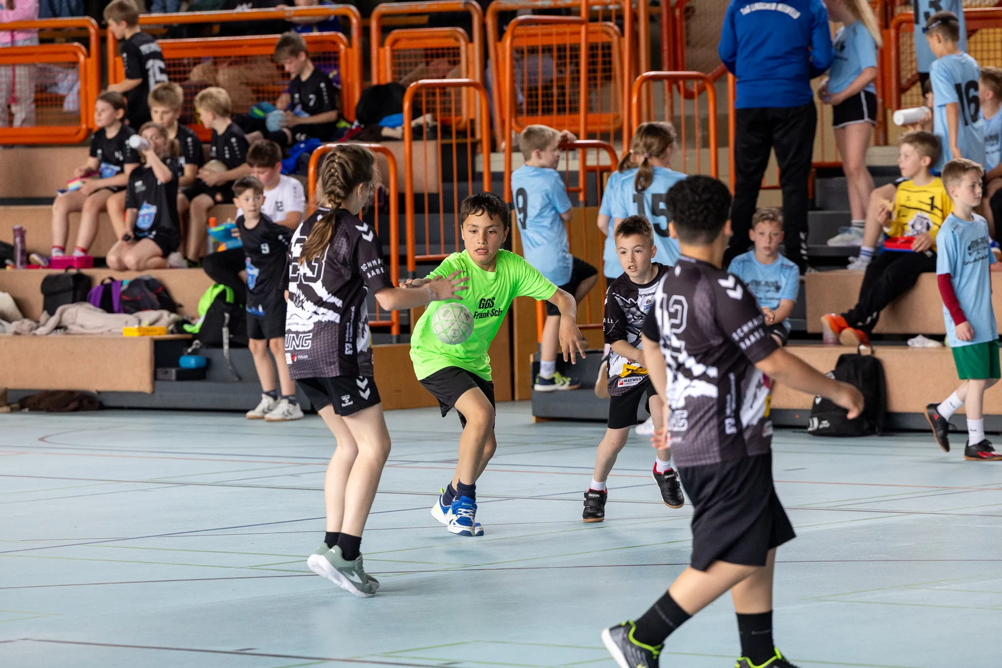 Handballspiel mit Kindern in Sporthalle: Ein Junge im grünen Trikot dribbelt den Ball, während zwei Gegenspielerinnen ihn verfolgen.