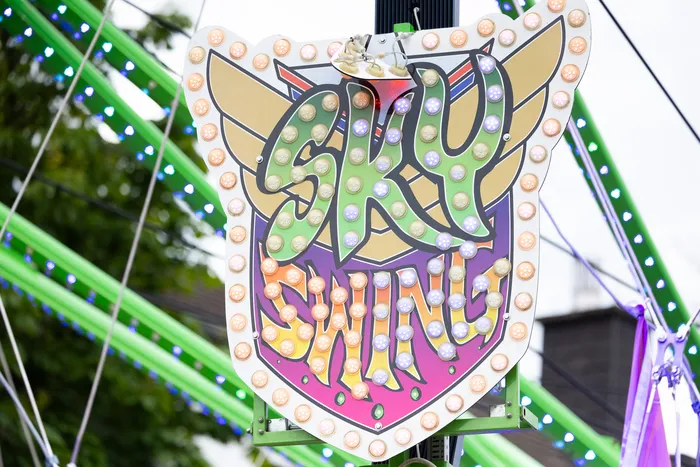 Leuchtendes Schild mit der Aufschrift „Sky Shine“ vor dem Hintergrund einer Kirmesattraktion mit grünen Lichtern.