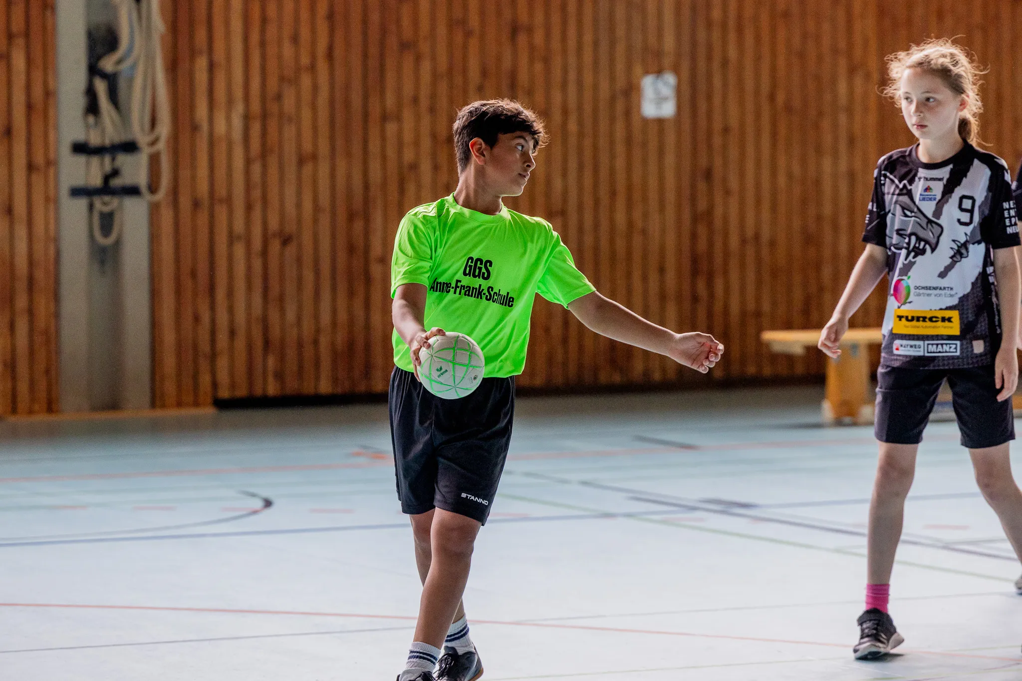 Zwei Jugendliche spielen Handball in einer Turnhalle. Der Spieler im grünen Trikot dribbelt den Ball, während sein Gegenspieler ihn beobachtet.