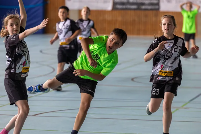 Ein Jugendlicher in einem leuchtend grünen Trikot dribbelt einen Ball während eines Handballspiels in einer Sporthalle. Zwei weitere Spielerinnen in schwarz-weißen Trikots stehen in der Nähe.