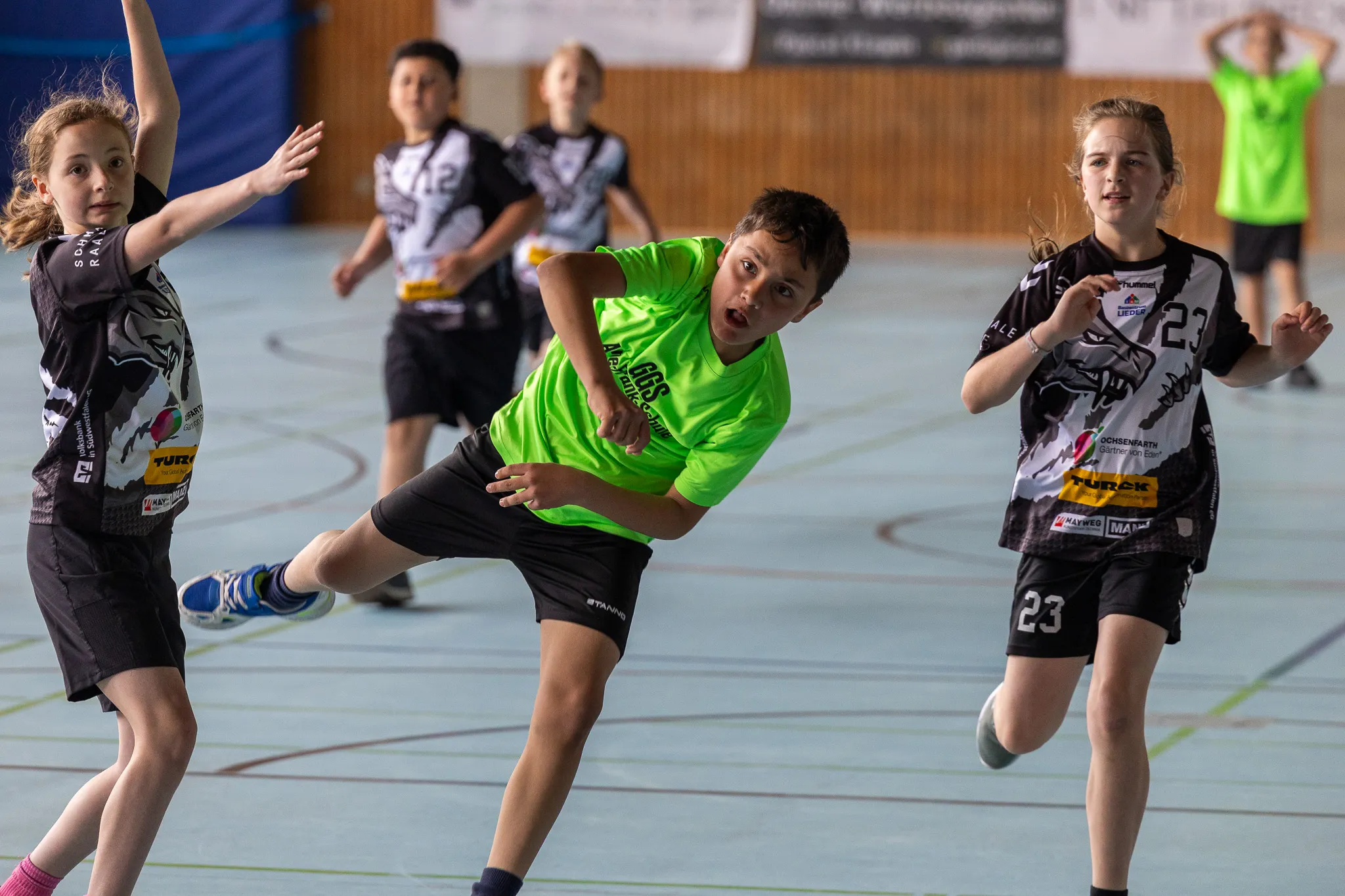 Ein Jugendlicher in einem leuchtend grünen Trikot dribbelt einen Ball während eines Handballspiels in einer Sporthalle. Zwei weitere Spielerinnen in schwarz-weißen Trikots stehen in der Nähe.