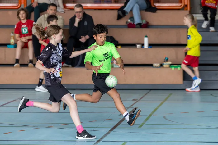 Zwei Kinder spielen Handball in einer Sporthalle. Ein Junge im grünen Trikot dribbelt den Ball, während ein Mädchen im schwarzen Trikot ihm entgegenkommt.