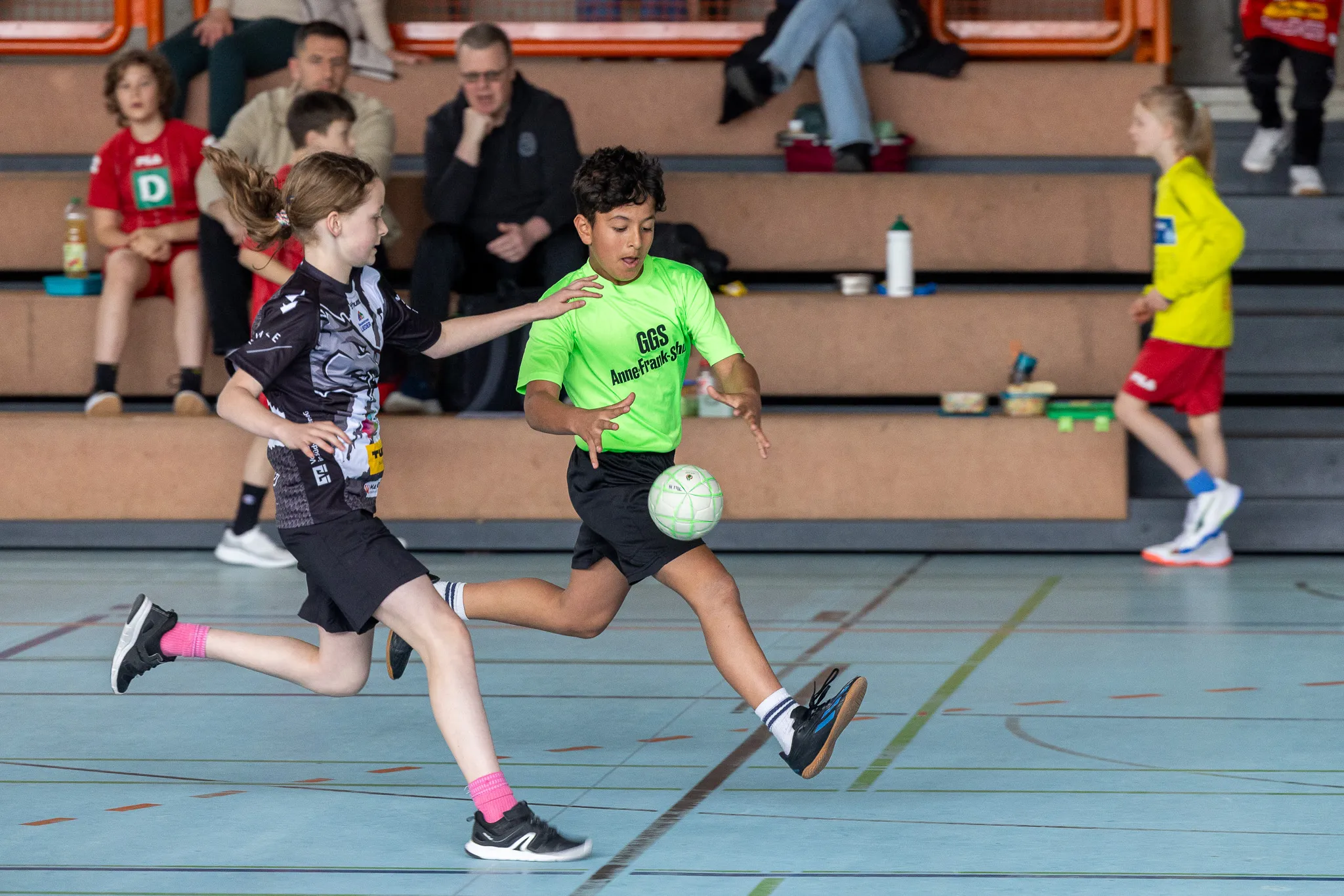 Zwei Kinder spielen Handball in einer Sporthalle. Ein Junge im grünen Trikot dribbelt den Ball, während ein Mädchen im schwarzen Trikot ihm entgegenkommt.