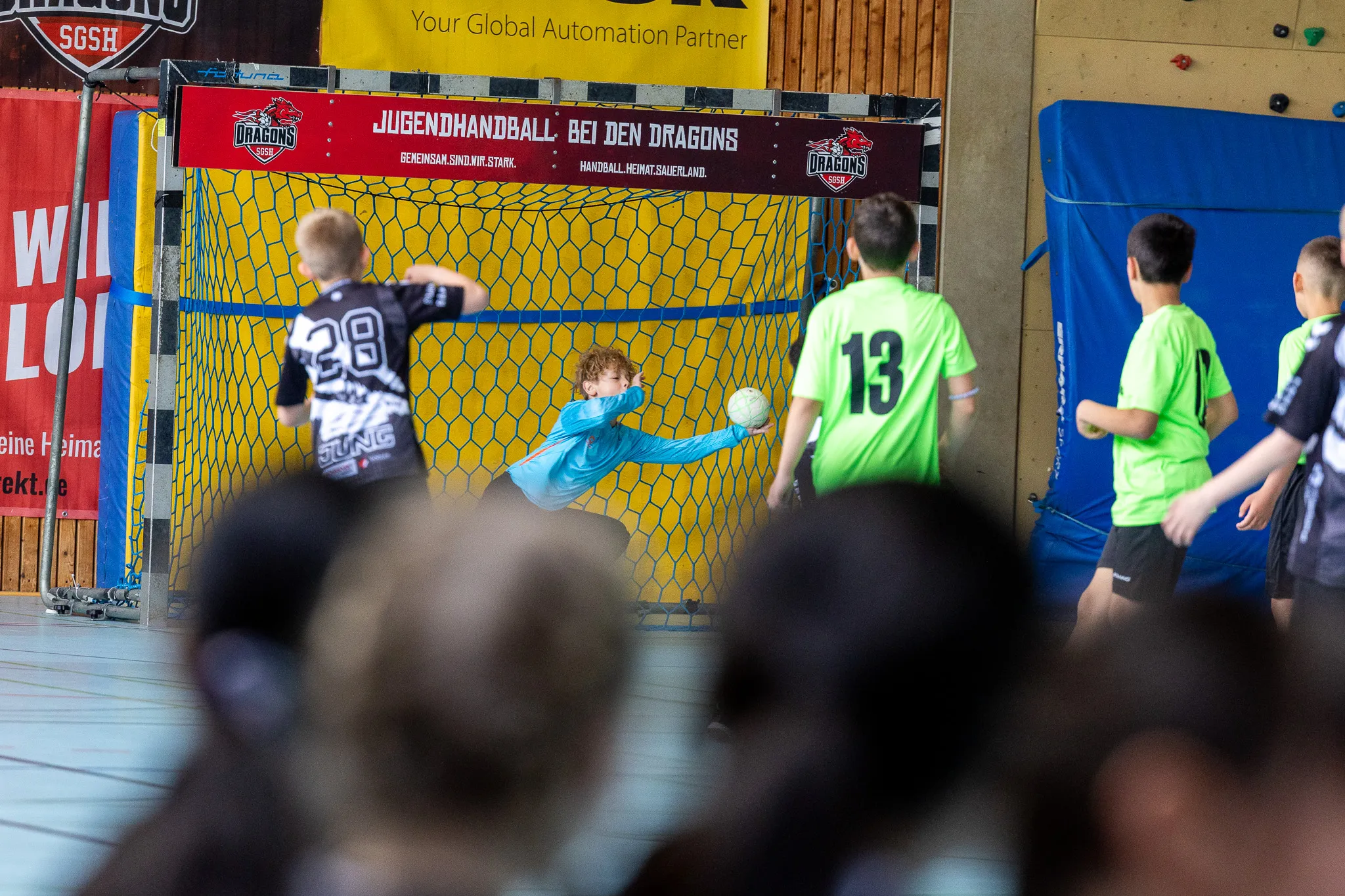 Handballspieler in grünen Trikots werfen in einem Hallensportraum auf das Tor. Im Hintergrund Banner mit dem Schriftzug „Jugendhandball bei den Dragons“.