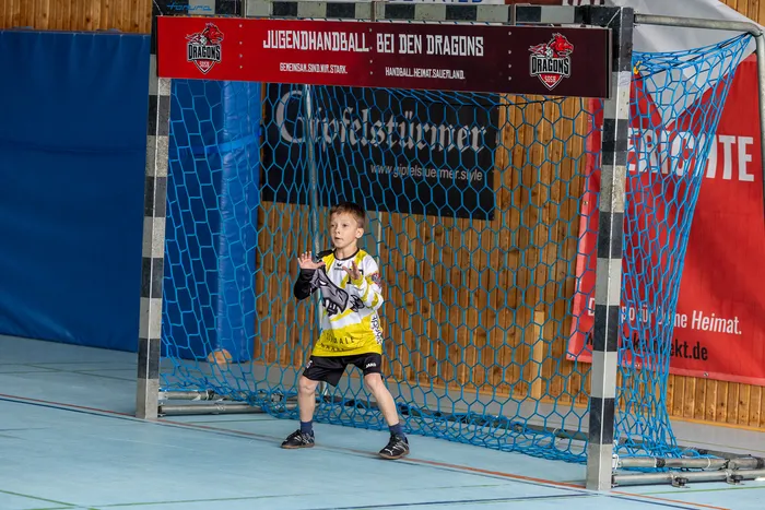 Junge im Handballtrikot steht vor einem blauen Tor mit roten Netzseiten und gestikuliert mit erhobener Hand.