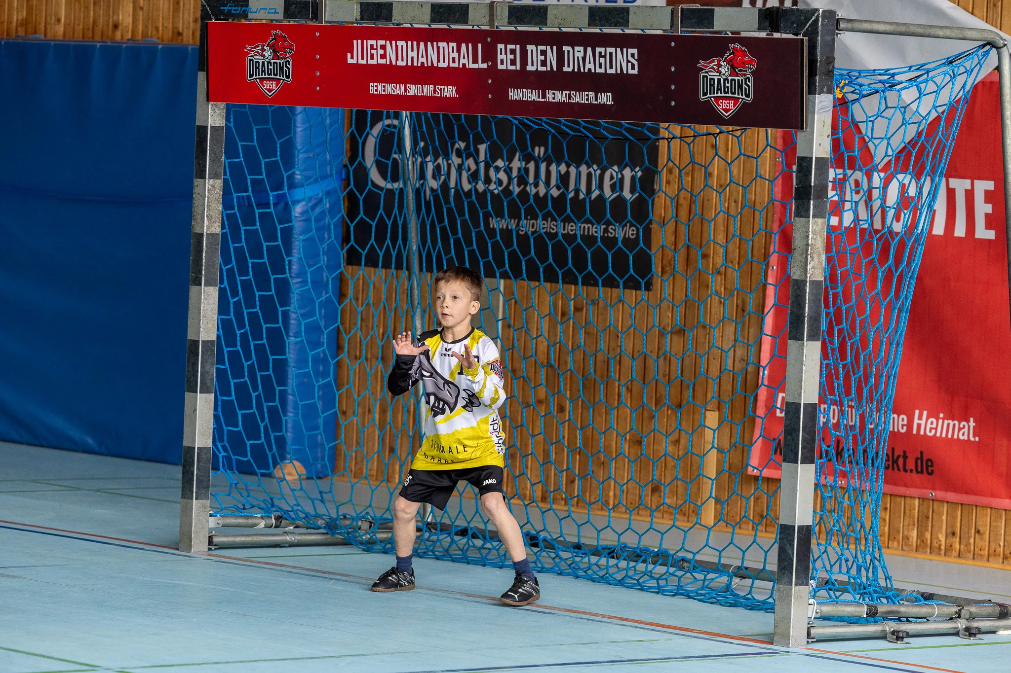 Junge im Handballtrikot steht vor einem blauen Tor mit roten Netzseiten und gestikuliert mit erhobener Hand.