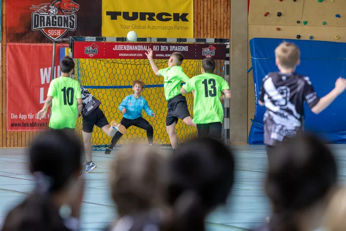 Handballtorwart in rotem Trikot und Shorts fängt den Ball vor zwei angreifenden Spielern in grünen Trikots ab.