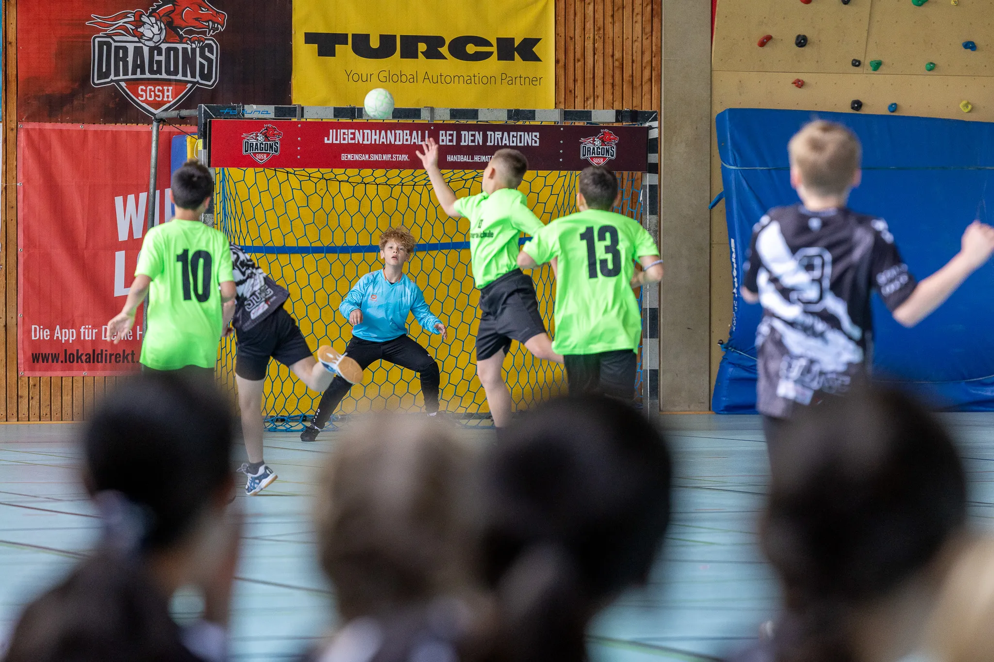 Handballtorwart in rotem Trikot und Shorts fängt den Ball vor zwei angreifenden Spielern in grünen Trikots ab.