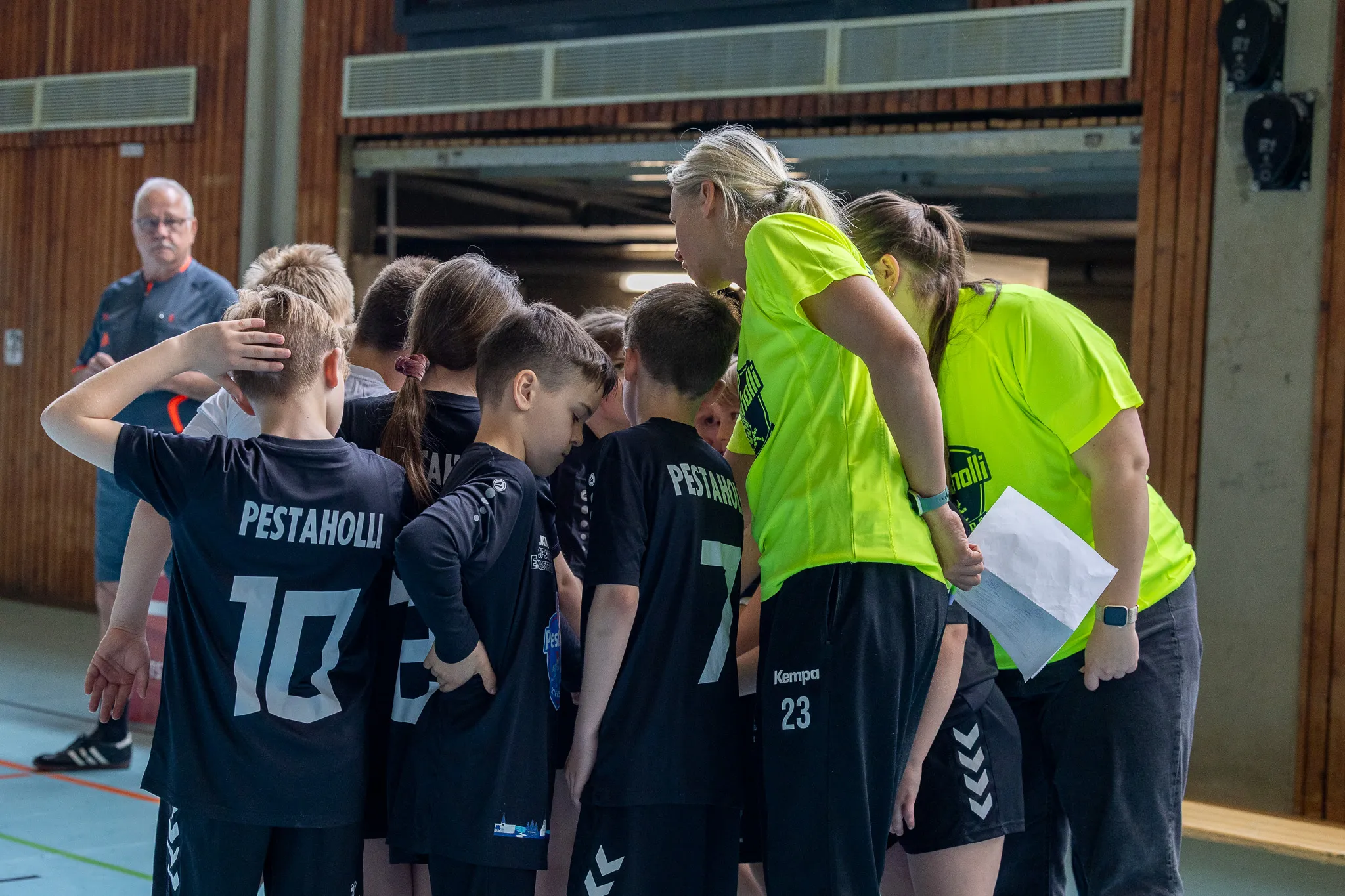 Trainerin spricht mit Jugendhandballmannschaft in schwarzer und neongelben Sportkleidung während einer Auszeit in einer Sporthalle.