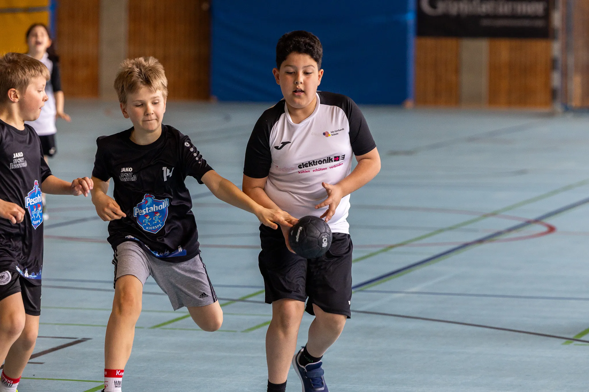Zwei Jungen beim Handballspiel in einer Sporthalle, ein dritter Spieler im Hintergrund. Die Trikots tragen den Aufdruck „Pestaholl“ und „JAKO“.
