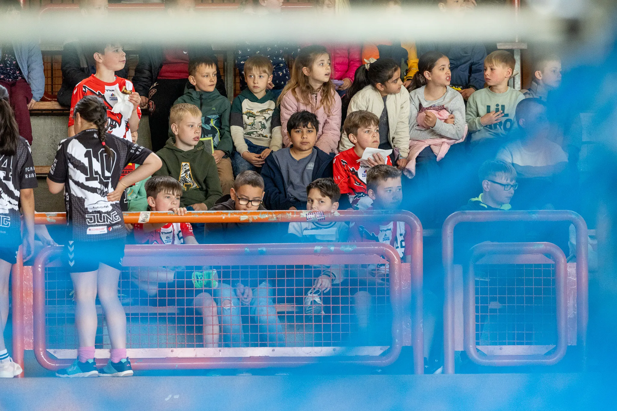 Zuschauer, hauptsächlich Kinder, beobachten ein Handballspiel hinter einem Zaun. Ein Spieler mit der Trikotnummer 10 steht im Vordergrund.
