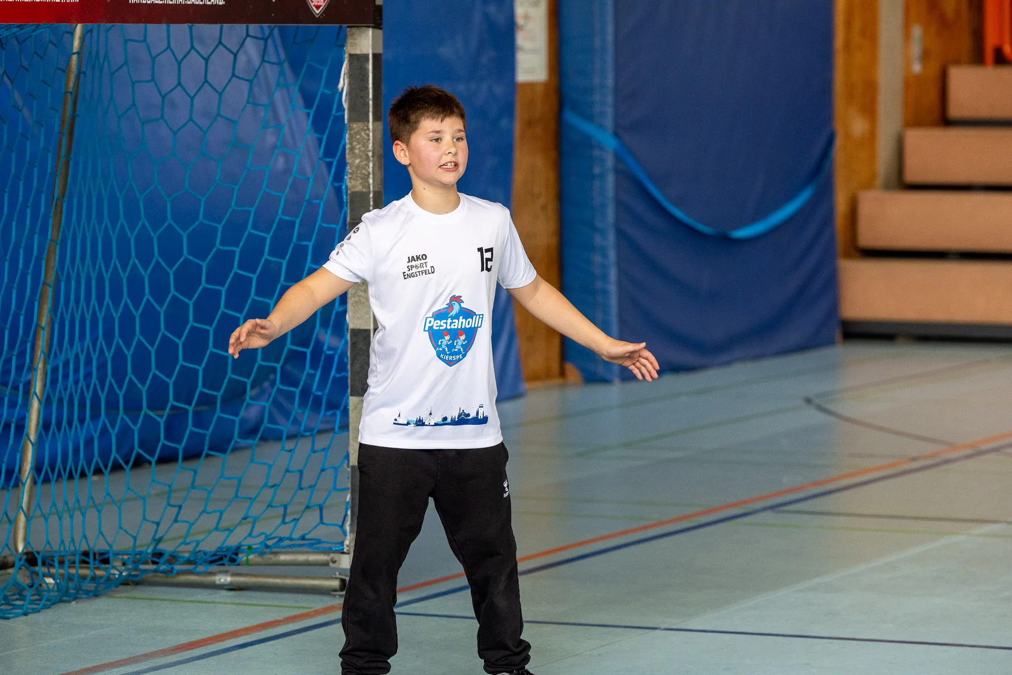 Junge im Handballtrikot mit der Nummer 12 steht in einer Sporthalle und streckt die Arme aus.