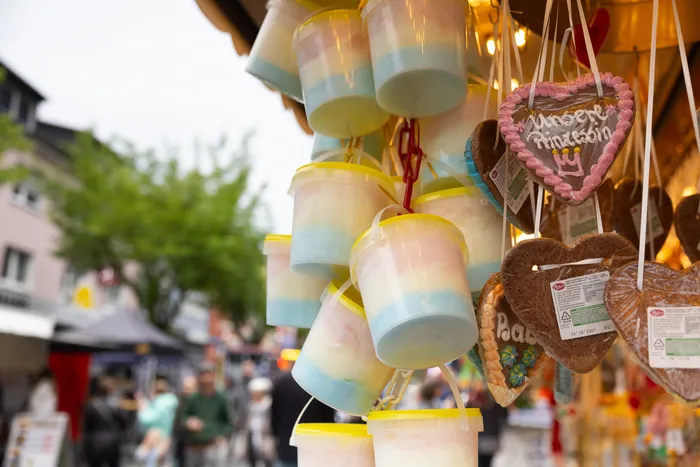 Zuckerwatte in Eimern und Lebkuchenherzen mit bunten Dekorationen hängen an einem Kirmesstand. Im Hintergrund sind Menschen zu sehen.