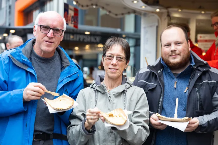 Drei Menschen essen auf der Kirmes Handbrote. Links ein älterer Mann mit Brille und blauer Jacke, in der Mitte eine Frau mit Brille und schwarzer Jacke, rechts ein jüngerer Mann mit Bart und grauer Jacke.
