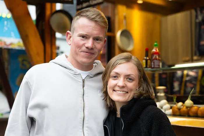 Ein Mann und eine Frau lächeln in die Kamera, im Hintergrund ein Marktstand mit Töpfen und Lebensmitteln.