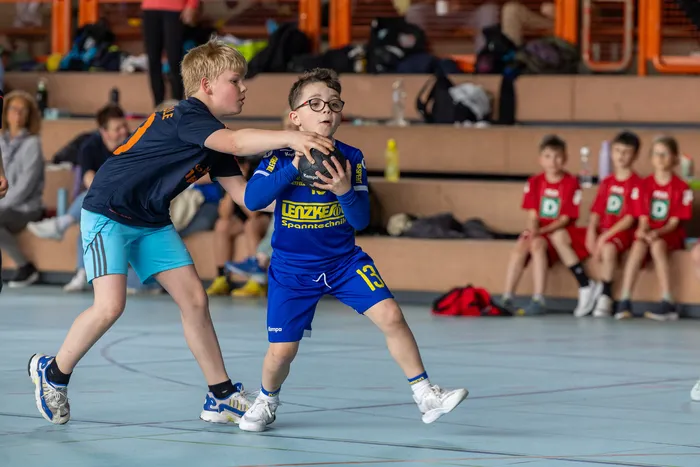 Zwei junge Handballspieler im Spiel: Ein Spieler in blauem Trikot mit Brille versucht, den Ball zu halten, während ein Gegner in dunklem Trikot ihn blockt.